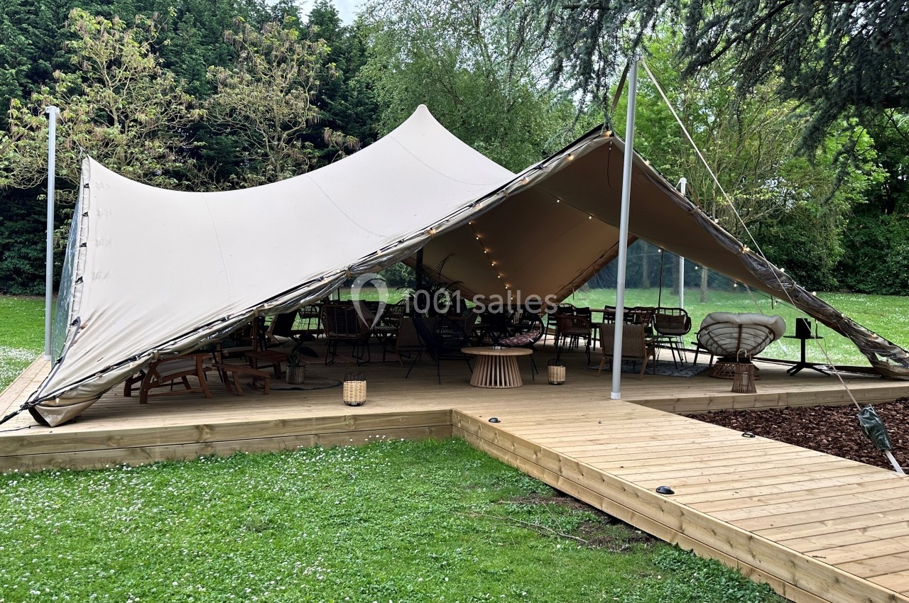 Tente beige montée sur une terrasse en bois, abritant des meubles de jardin dans un espace verdoyant.
