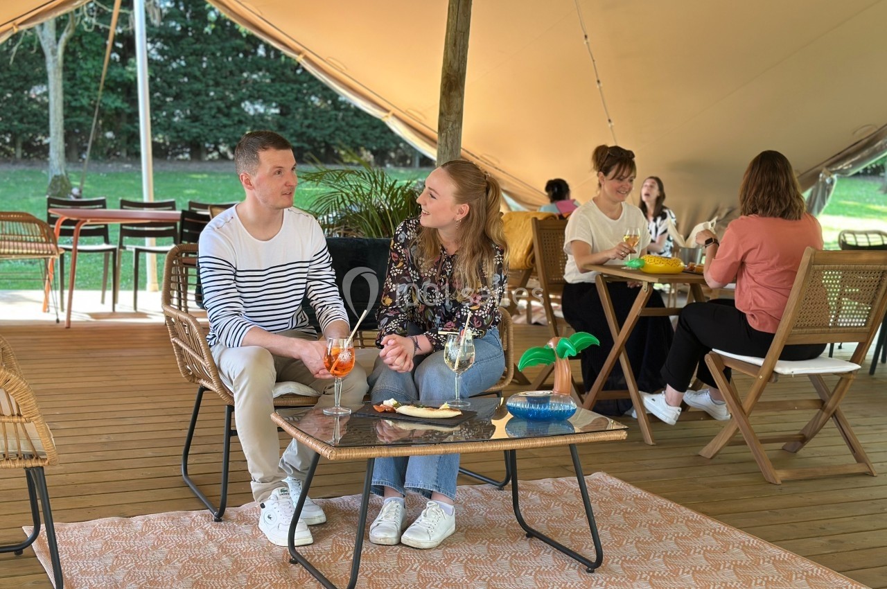 Deux personnes discutent autour d'une table avec des boissons sous une tente, d'autres personnes assises en arrière-plan.