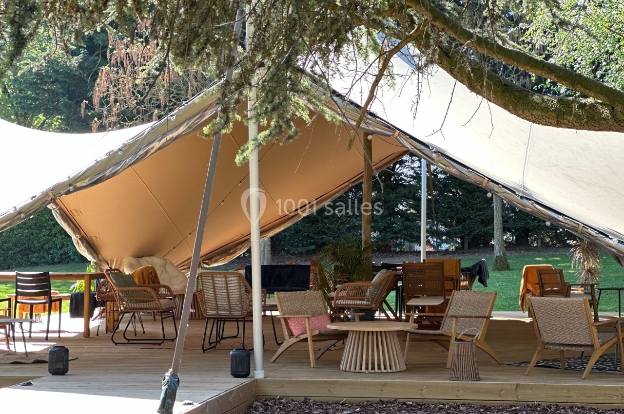 Espace extérieur aménagé sous une tente avec des meubles en bois et rotin, entouré de verdure.