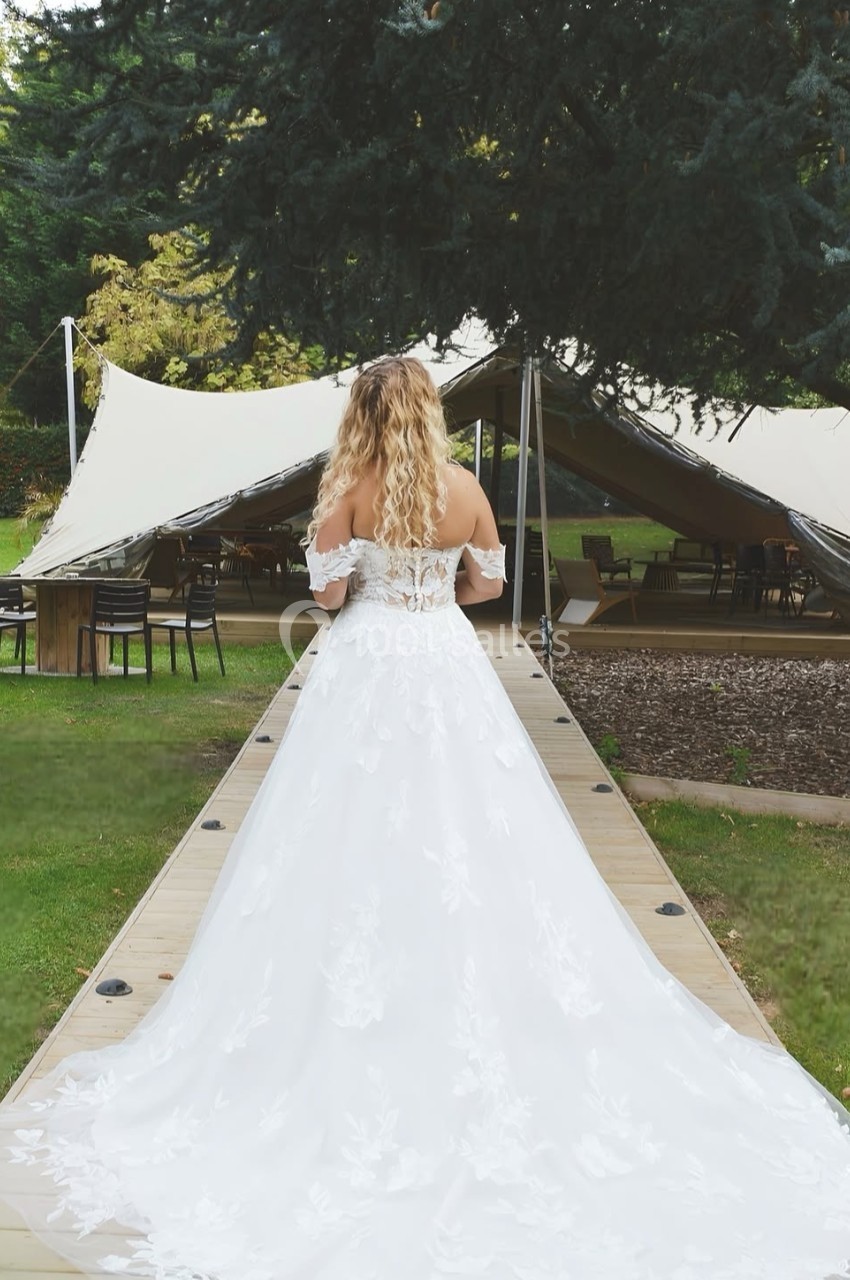 Location salle Noyelles-Godault (Pas-de-Calais) - Novotel Lens Noyelles #5 Femme en robe de mariée blanche vue de dos, marchant sur une passerelle en bois vers une tente dans un jardin.