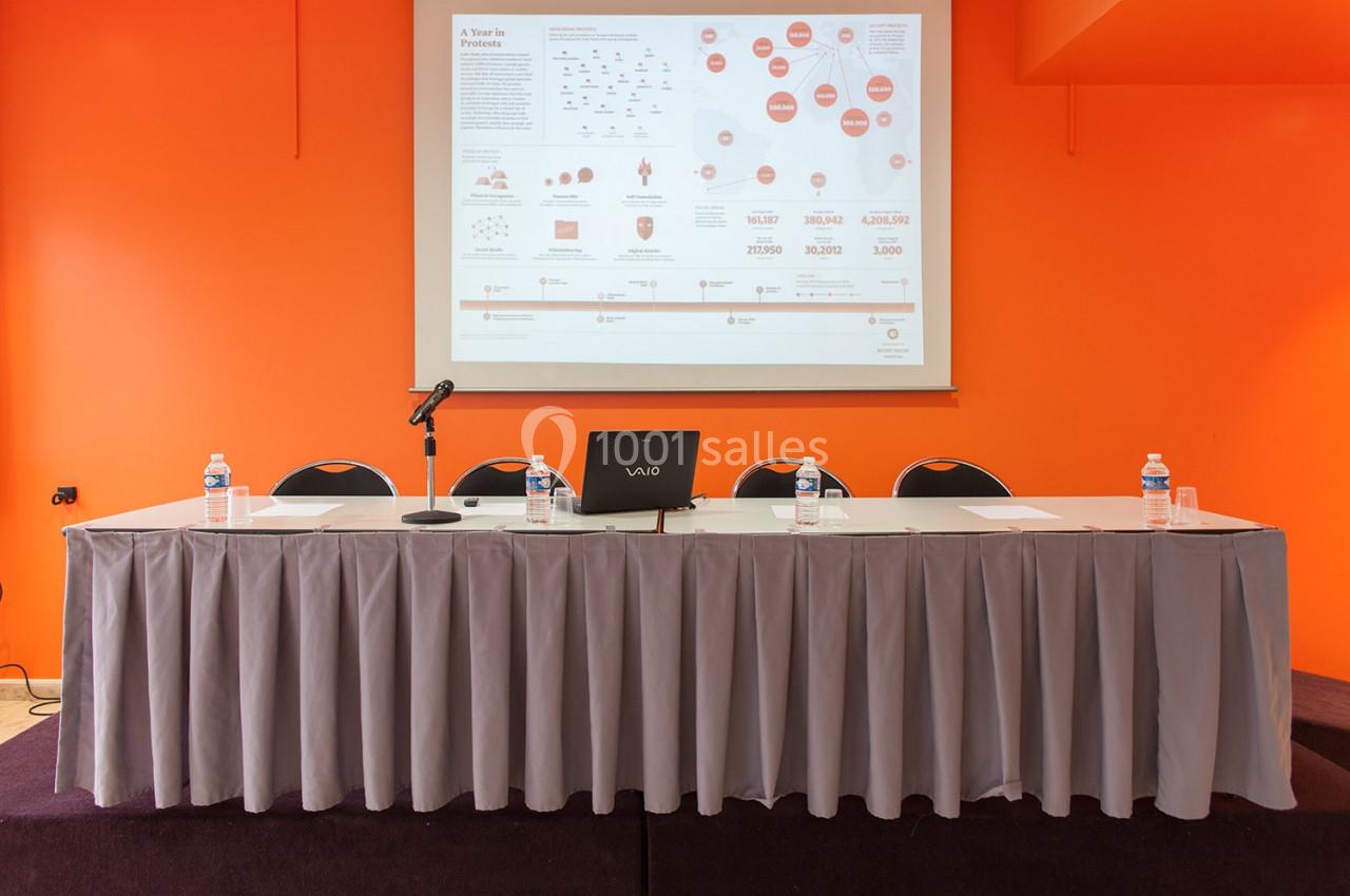 Table avec micro, ordinateur portable et bouteilles d'eau devant un écran de présentation dans une salle orange.