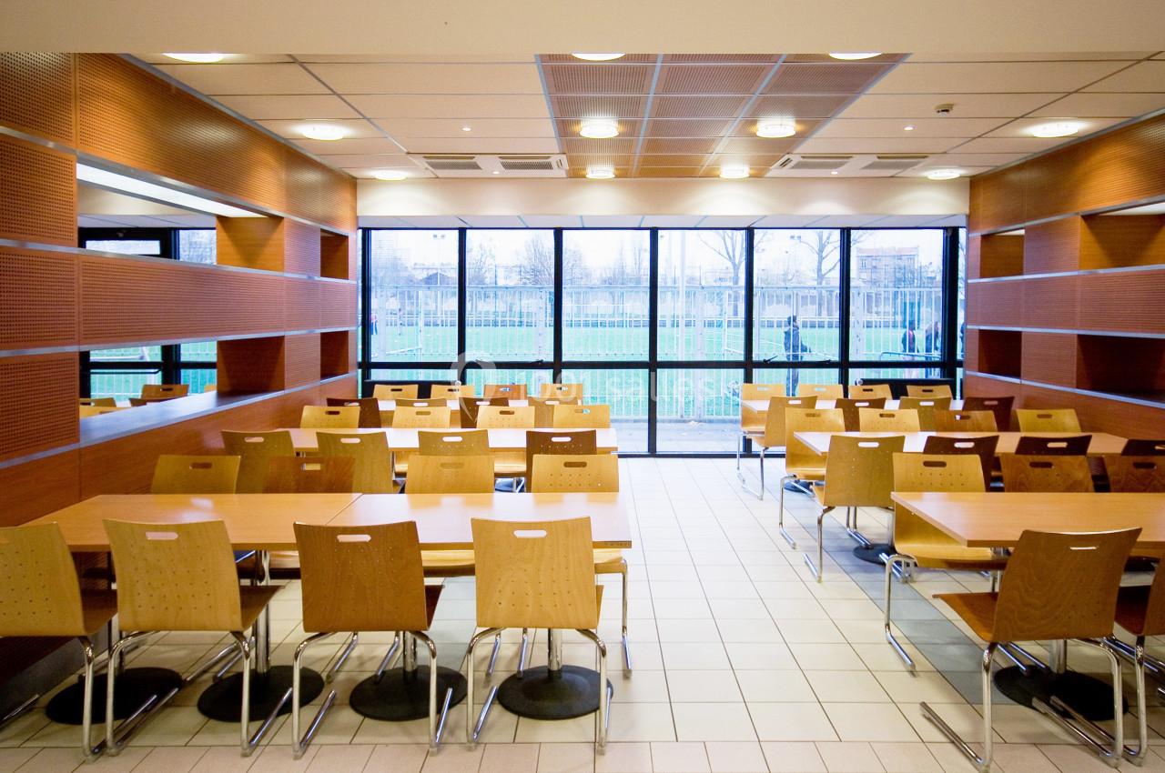 Salle de restauration lumineuse avec tables et chaises en bois, grandes fenêtres donnant sur un espace extérieur.