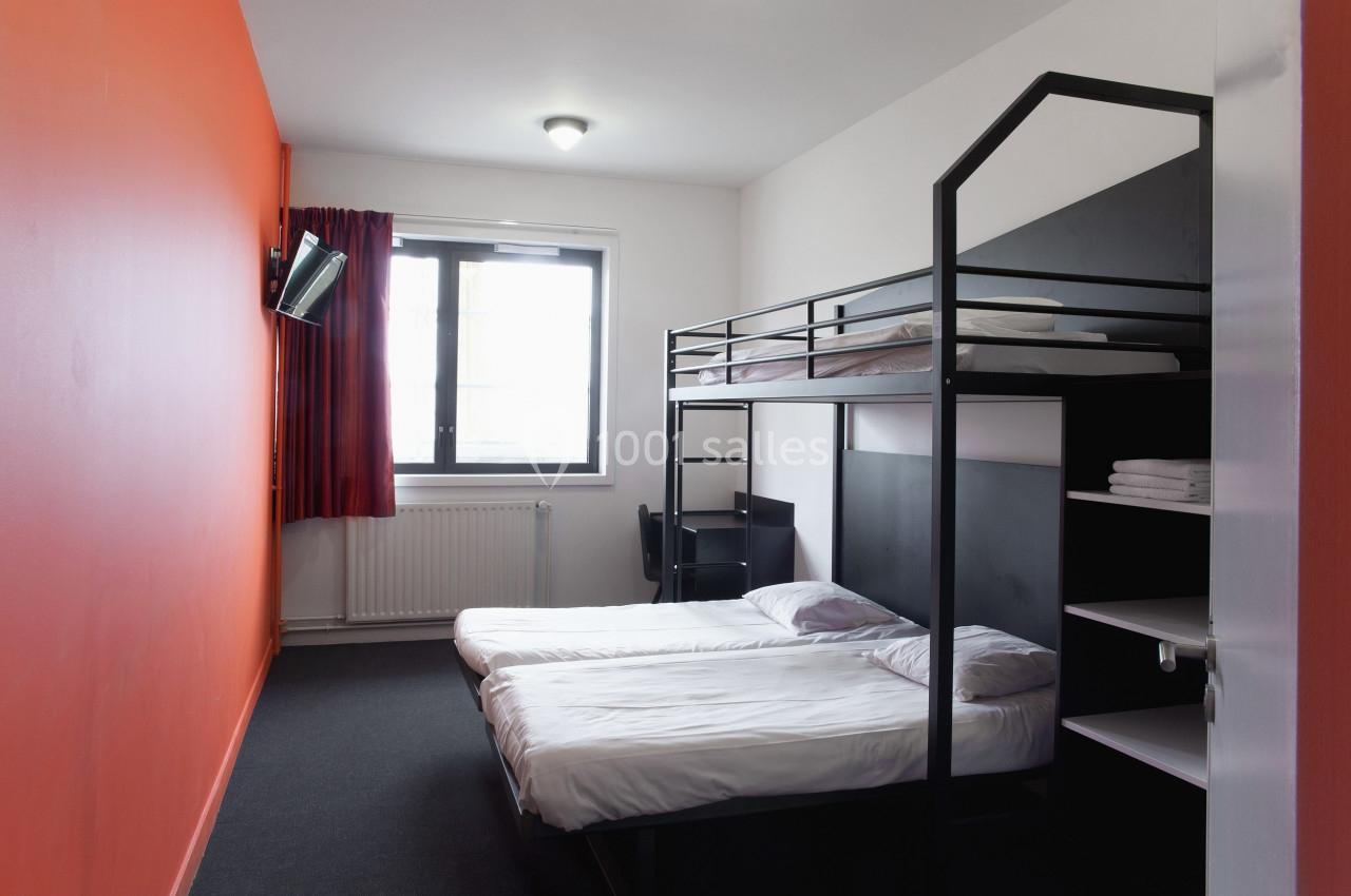 Chambre avec lits superposés et lit simple, mur orange, fenêtre avec rideaux rouges, télévision et étagères.