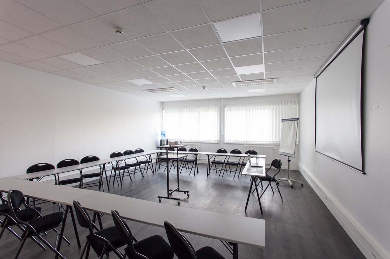 Salle de réunion lumineuse avec tables disposées en U, chaises noires, écran de projection et tableau de conférence.