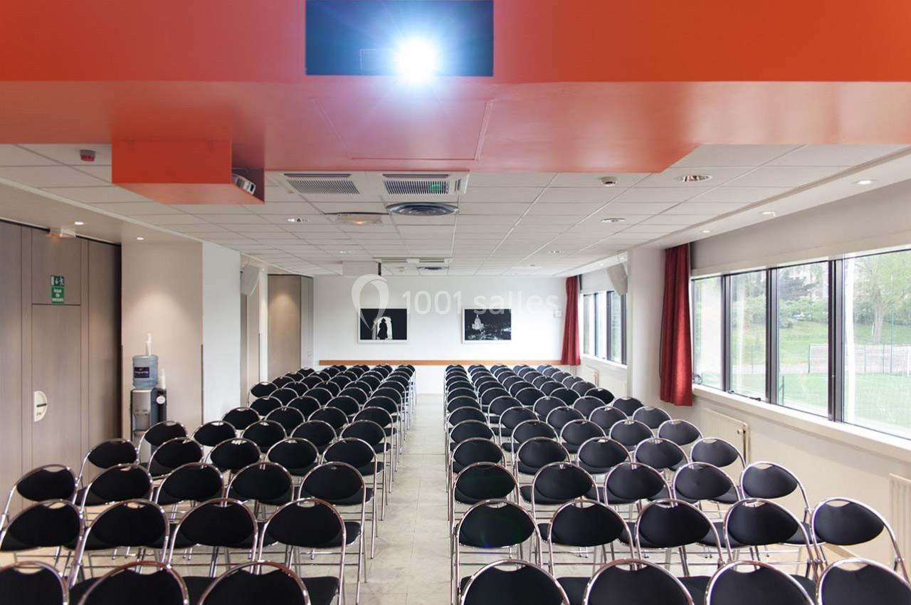 Salle de conférence lumineuse avec des rangées de chaises noires alignées et un projecteur allumé au plafond.
