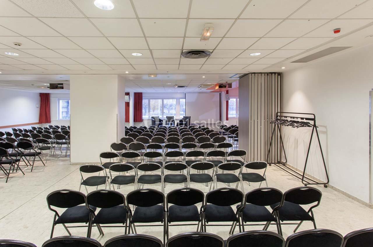 Salle de conférence avec des rangées de chaises noires alignées face à un écran de projection et des fenêtres.