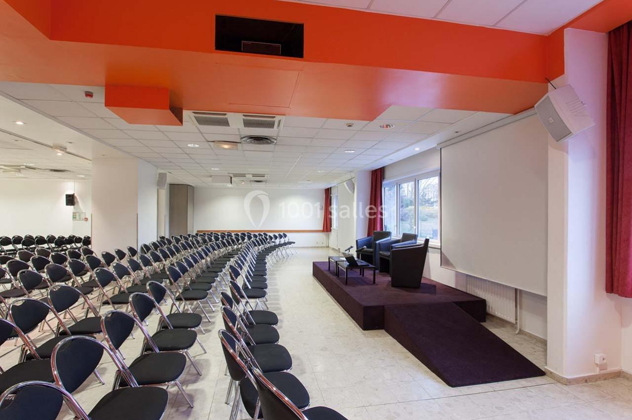 Salle de conférence lumineuse avec rangées de chaises noires, estrade équipée de fauteuils et écran de projection.