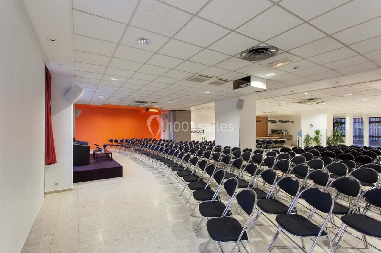 Salle de conférence vide avec des rangées de chaises noires pliantes, une scène et un mur orange au fond.