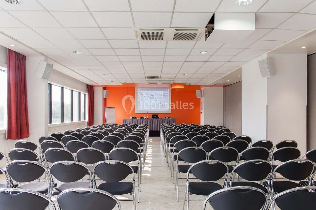 Salle de conférence vide avec des rangées de chaises noires, un écran de projection et un mur orange en arrière-plan.