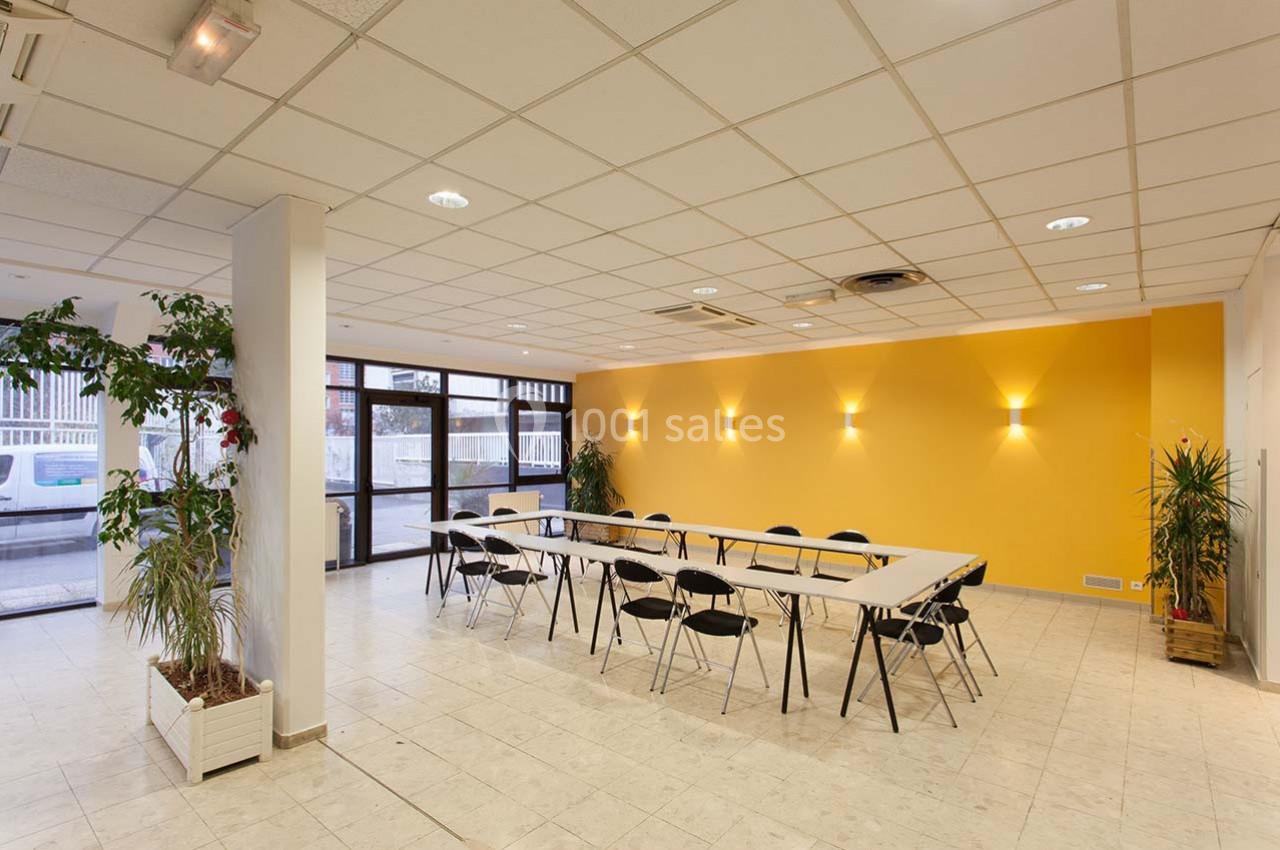 Salle de réunion lumineuse avec murs jaunes, grandes fenêtres, tables disposées en rectangle et chaises noires pliantes.