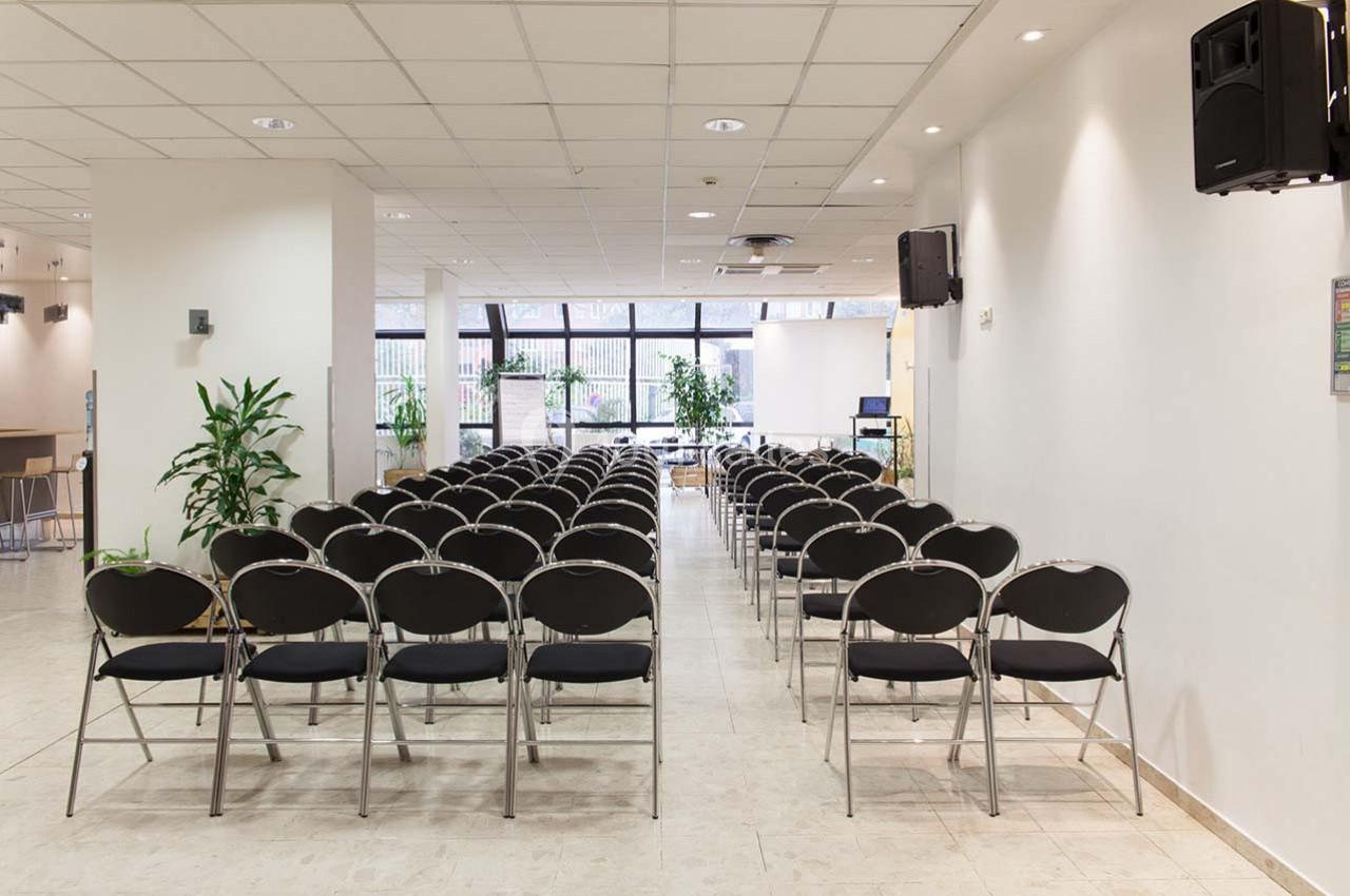 Salle de conférence lumineuse avec des rangées de chaises noires alignées face à un espace de présentation.