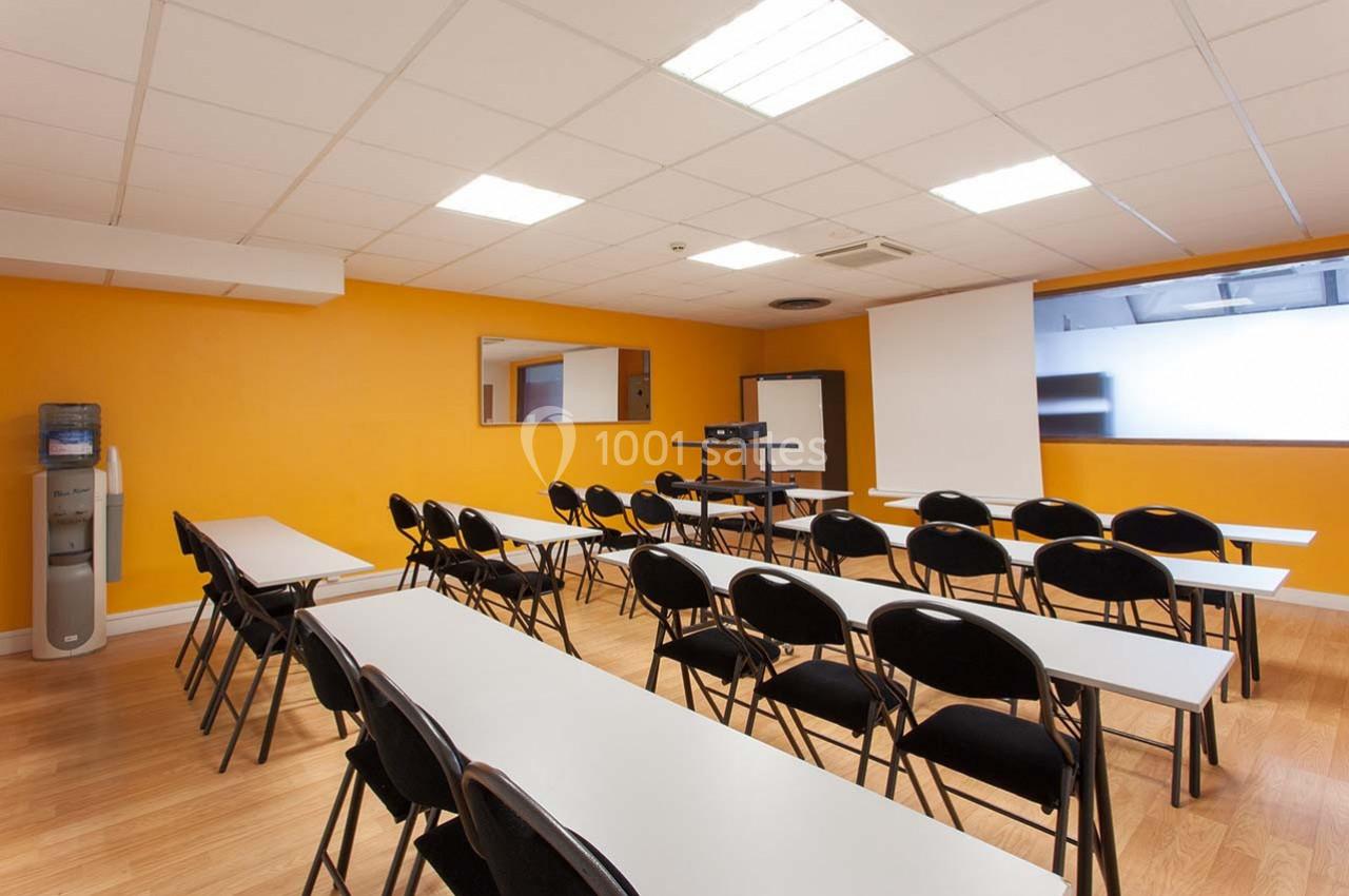 Salle de formation avec murs orange, tables blanches alignées, chaises noires et un écran de projection.