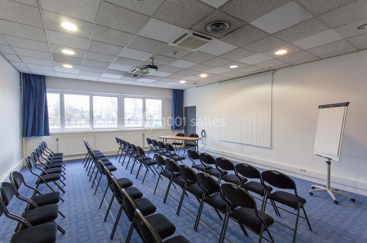 Salle de réunion vide avec rangées de chaises noires, écran de projection, tableau blanc et grandes fenêtres.