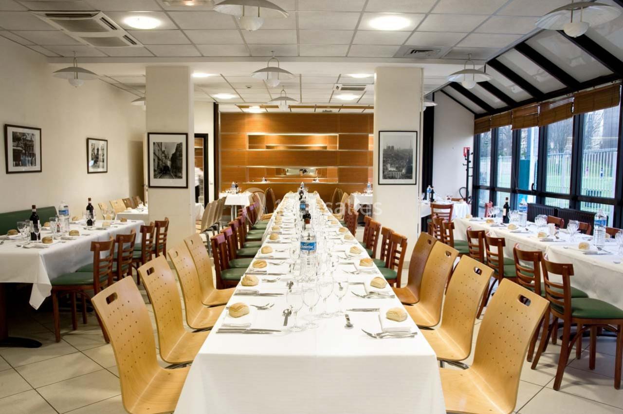 Salle de restaurant avec de longues tables dressées, chaises en bois, nappes blanches et lumière naturelle.