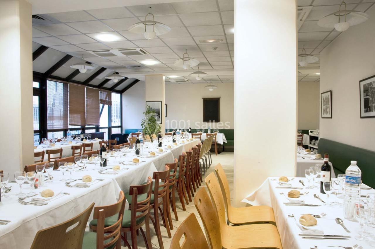 Salle de restaurant lumineuse avec tables dressées, nappes blanches, chaises en bois et grandes fenêtres.