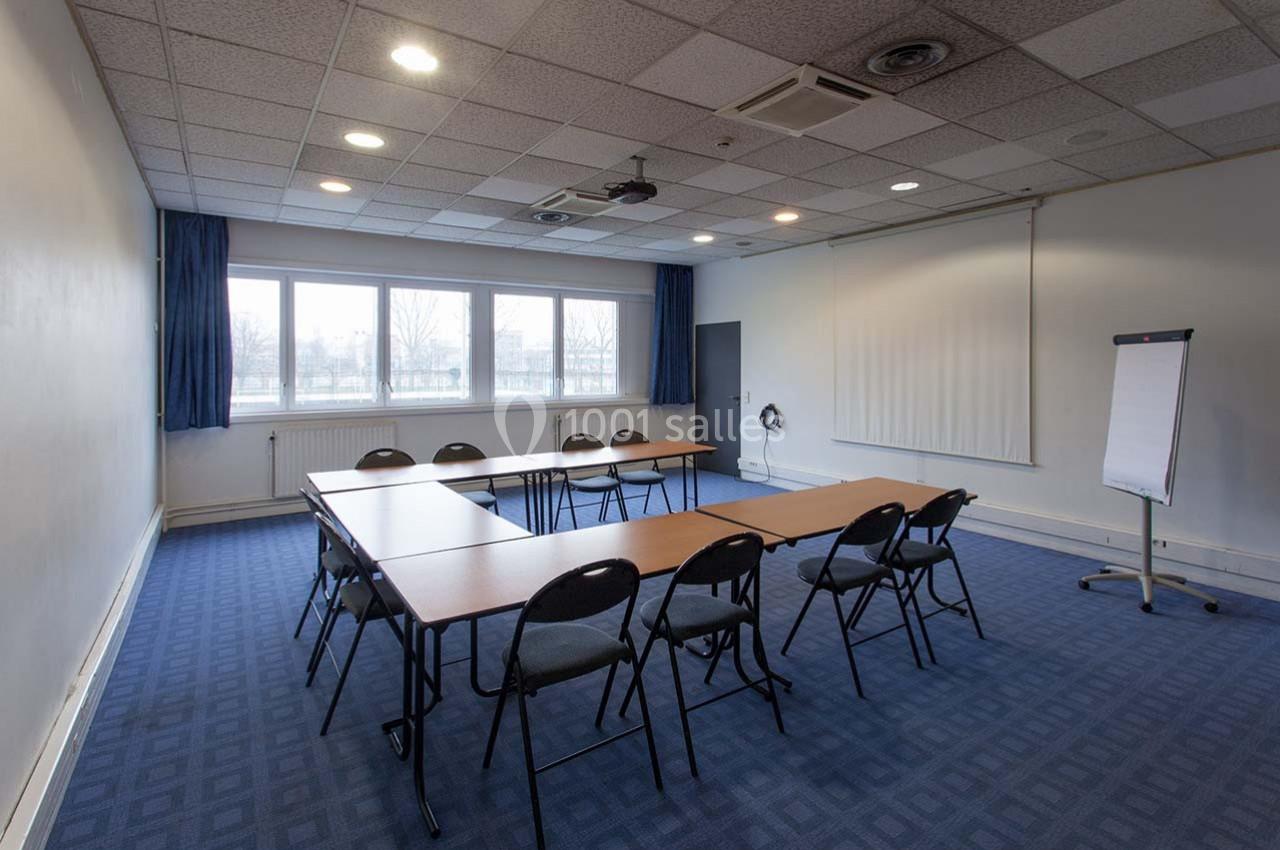 Salle de réunion avec tables disposées en U, chaises, paperboard, projecteur au plafond et fenêtres laissant entrer la…