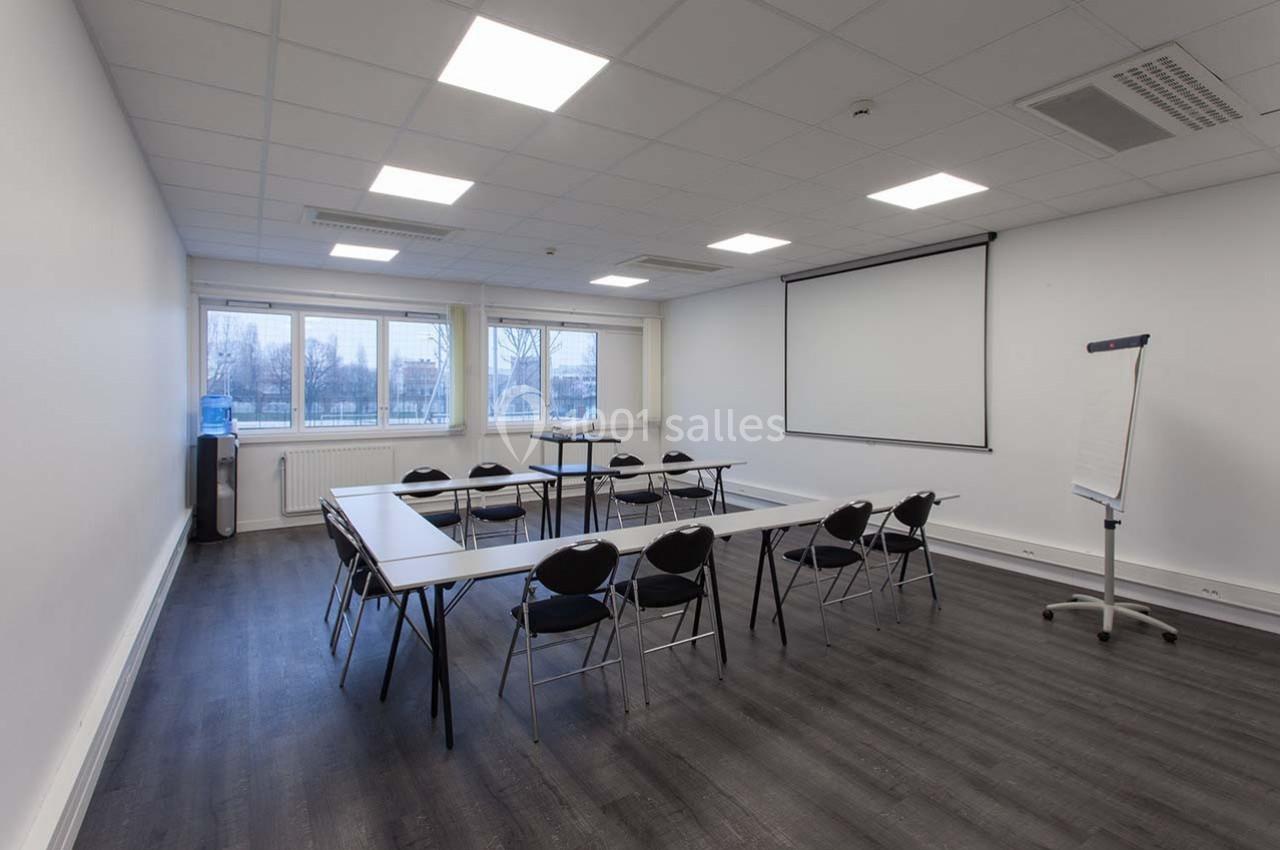 Salle de réunion lumineuse avec tables disposées en U, chaises, tableau blanc et écran de projection.