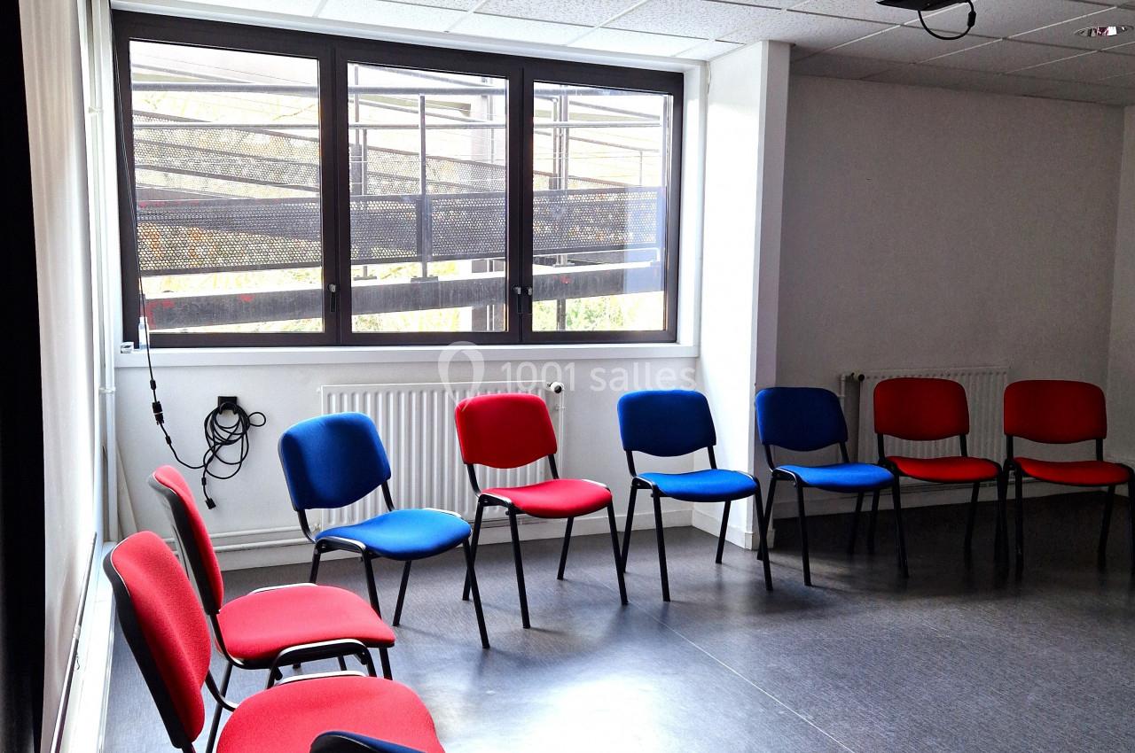 Salle de réunion vide avec des chaises rouges et bleues disposées en cercle, éclairée par une lumière naturelle.