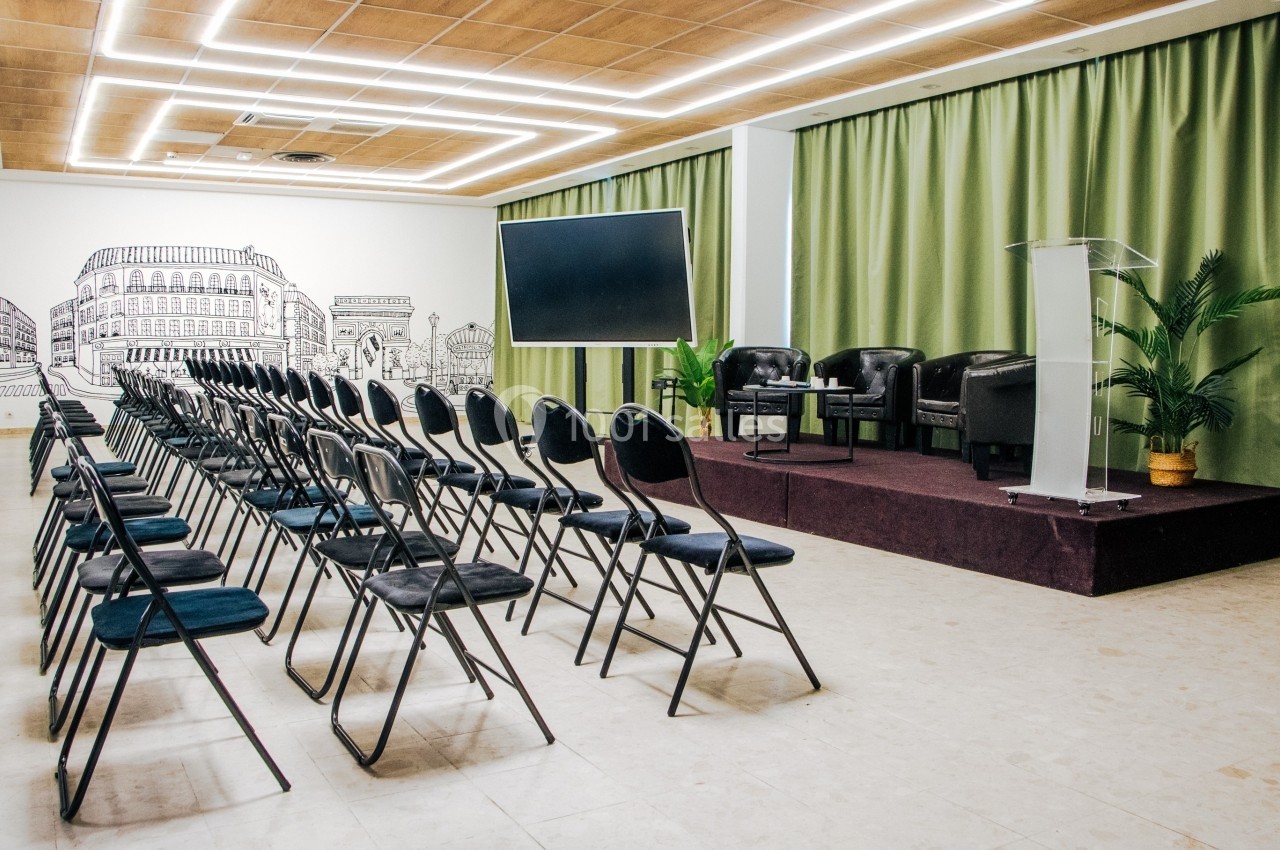 Salle de conférence avec rangées de chaises noires, une estrade équipée de fauteuils, un pupitre et un écran.