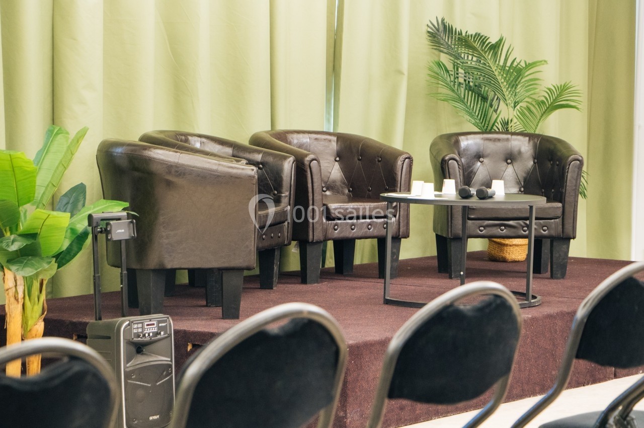 Quatre fauteuils en cuir disposés sur une estrade avec une table basse, des micros et des plantes en arrière-plan.