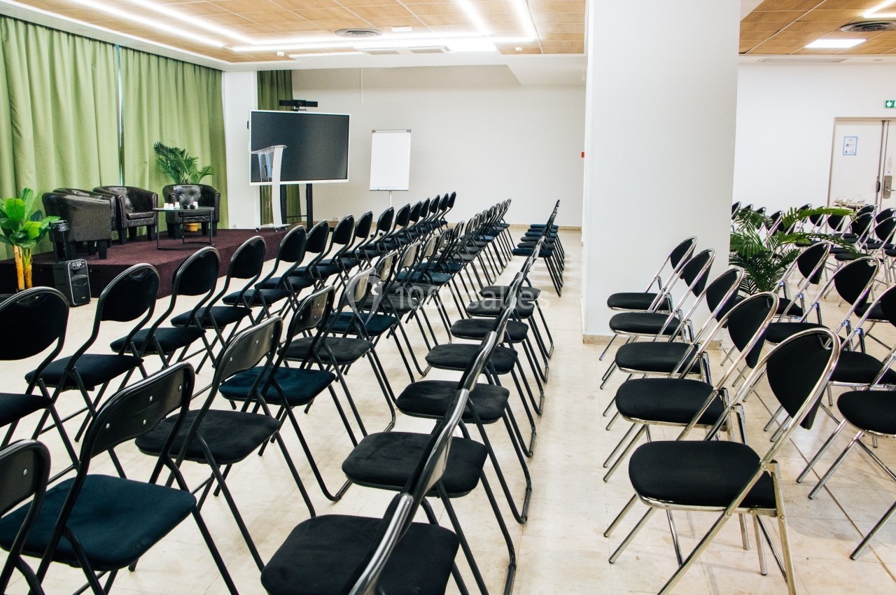 Salle de conférence avec des rangées de chaises noires, une scène équipée de fauteuils et un écran de présentation.