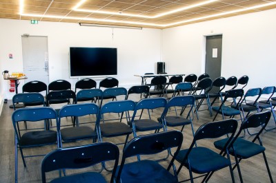 Chambre lumineuse avec deux lits simples, une table, une lampe, une fenêtre avec rideaux et une étagère grise.