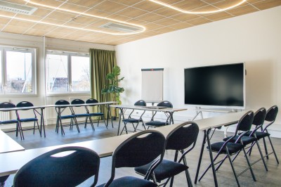 Chambre lumineuse avec deux lits simples, une table, une lampe, une fenêtre avec rideaux et une étagère grise.