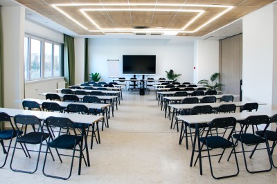 Chambre lumineuse avec deux lits simples, une table, une lampe, une fenêtre avec rideaux et une étagère grise.