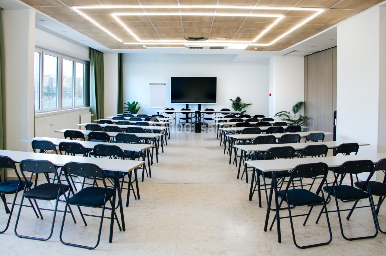 Salle de conférence lumineuse avec des rangées de tables et chaises noires, un écran et un tableau blanc à l'avant.
