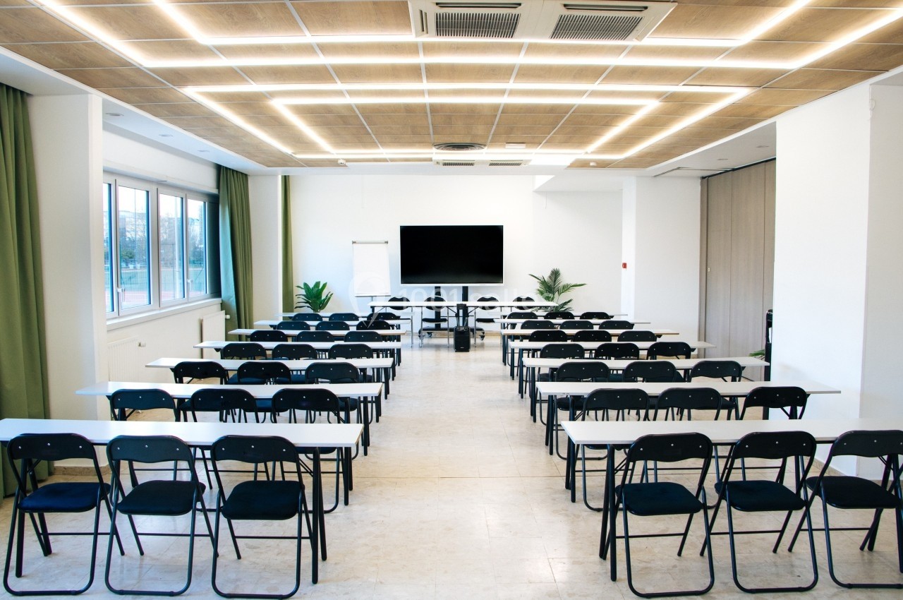 Salle de conférence lumineuse avec des rangées de tables et chaises noires, un écran et un tableau blanc à l'avant.