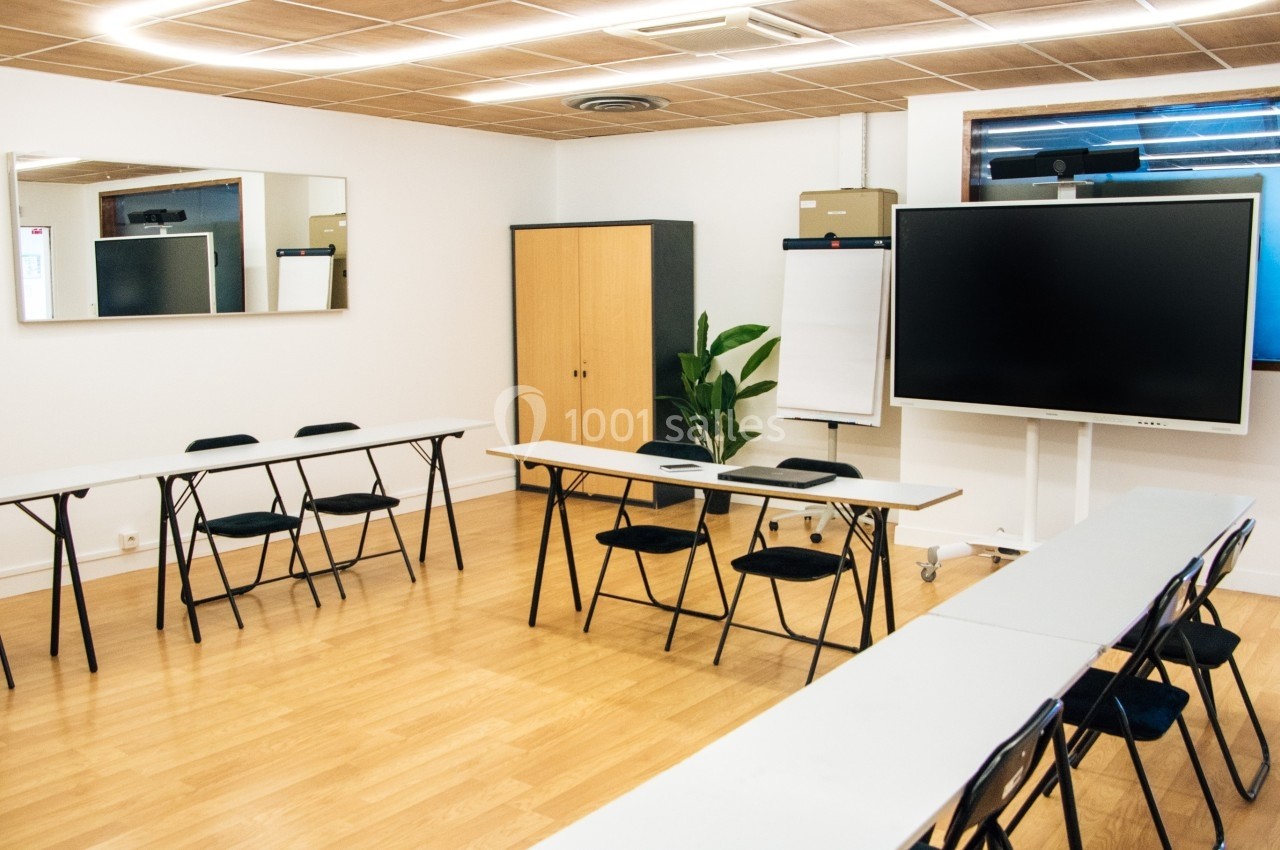Salle de réunion lumineuse avec tables disposées en U, tableau interactif, paperboard et armoire en arrière-plan.