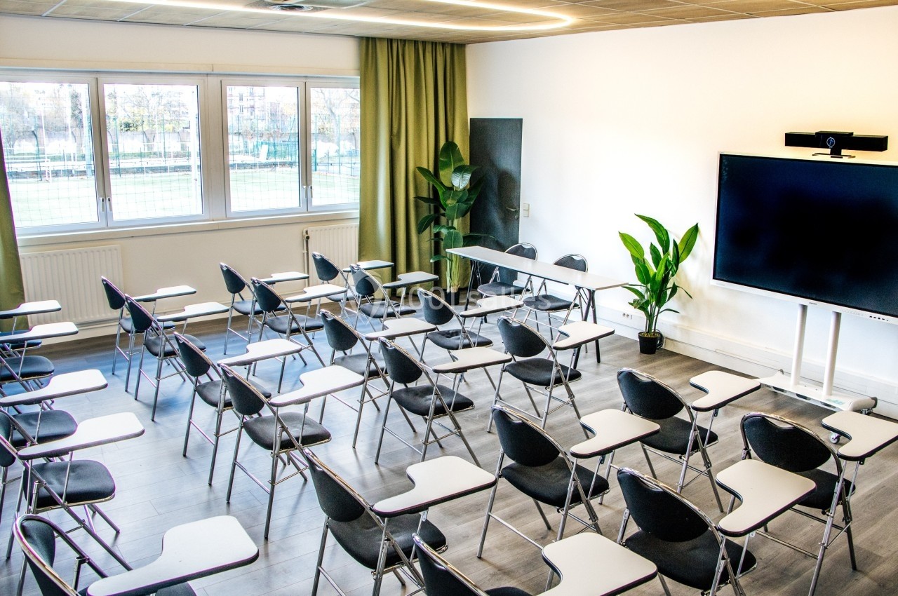 Salle de classe moderne avec des chaises équipées de tablettes, un écran interactif et des plantes en arrière-plan.
