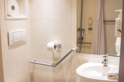 Chambre lumineuse avec deux lits simples, une table, une lampe, une fenêtre avec rideaux et une étagère grise.