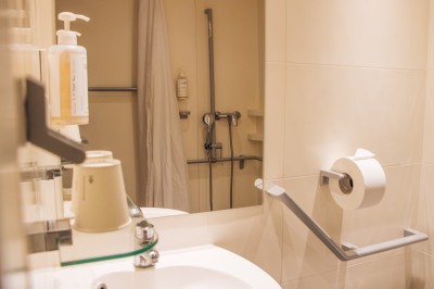 Chambre lumineuse avec deux lits simples, une table, une lampe, une fenêtre avec rideaux et une étagère grise.