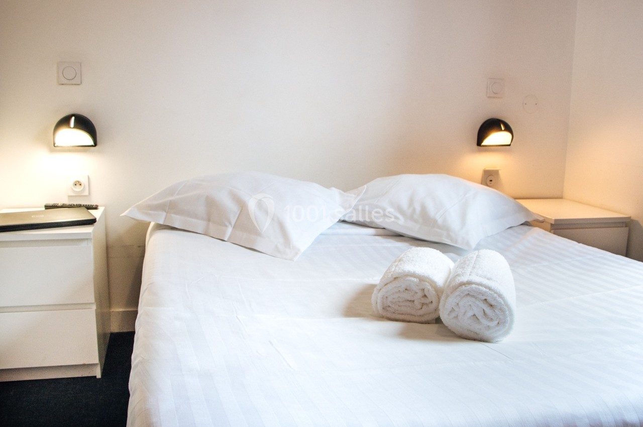 Lit double avec draps blancs, deux oreillers, deux serviettes roulées et lampes murales allumées dans une chambre simple.