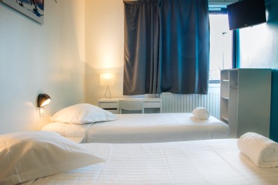 Chambre lumineuse avec deux lits simples, une table, une lampe, une fenêtre avec rideaux et une étagère grise.