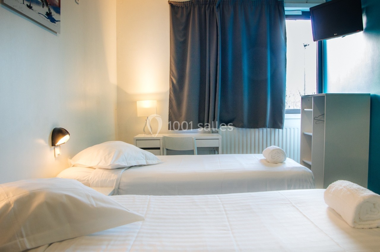 Chambre lumineuse avec deux lits simples, une table, une lampe, une fenêtre avec rideaux et une étagère grise.