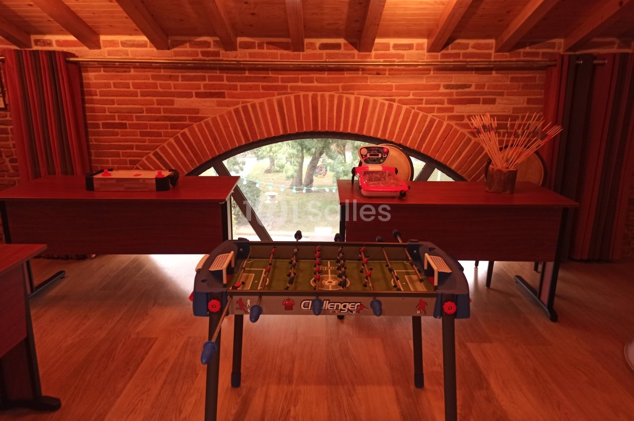 Table de baby-foot dans une pièce avec murs en briques, plafond en bois et grande fenêtre en arc donnant sur l'extérieur.