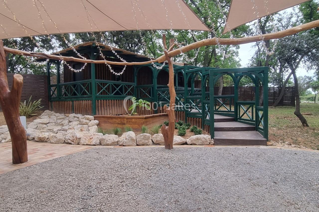Terrasse en bois avec balustrades vertes, entourée d'arbres et de voiles d'ombrage, sur un sol en gravier et pavés.