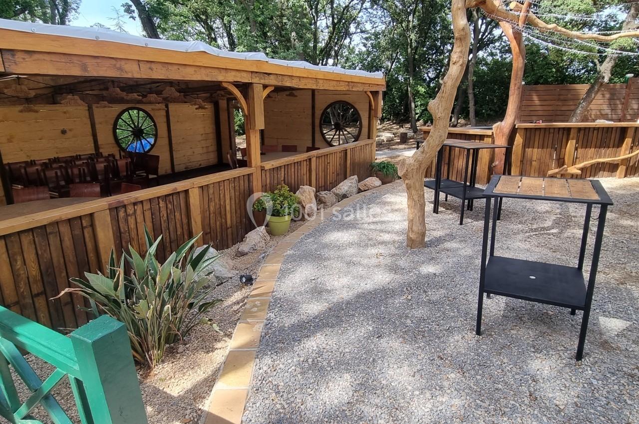 Espace extérieur avec terrasse en bois, tables noires, plantes et arbres, entouré d'une clôture en bois.