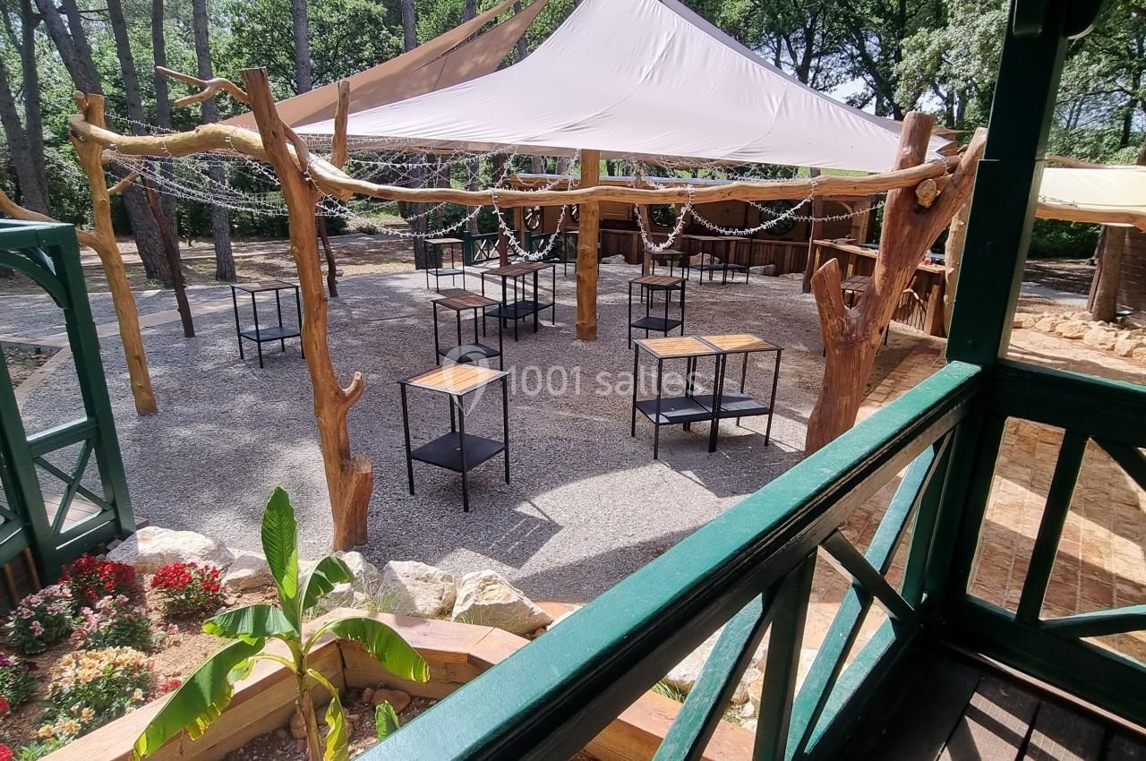 Espace extérieur avec tables et chaises sous une toile d'ombrage, entouré de végétation et de structures en bois.