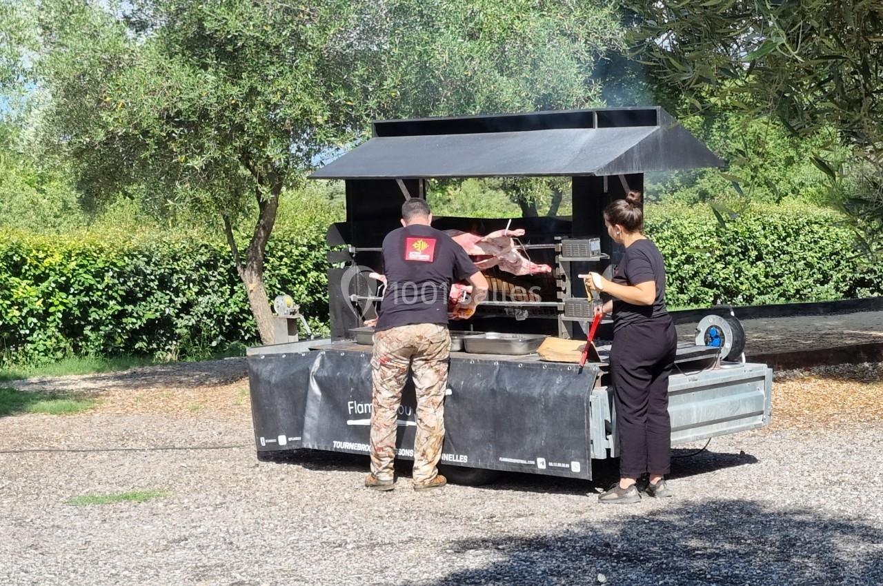 Deux personnes cuisinent un cochon entier sur une rôtissoire mobile en plein air, entourées de végétation.