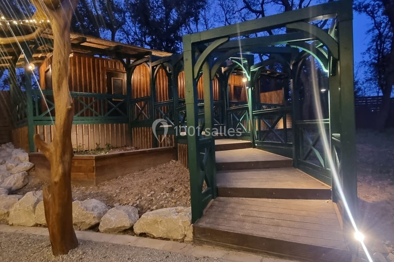 Passerelle en bois avec structure métallique verte, éclairée par des lumières, menant à une terrasse en soirée.