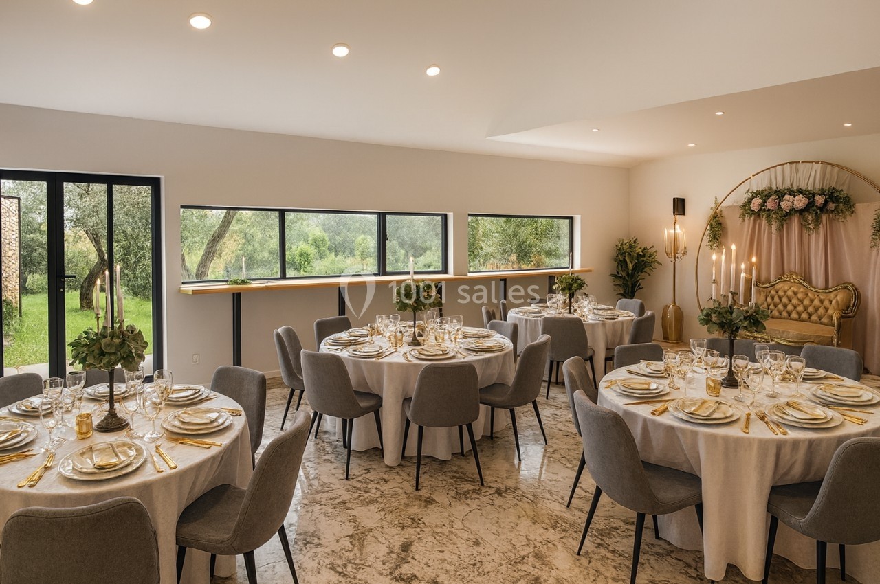 Salle de réception lumineuse avec tables rondes dressées, chaises grises et vue sur un jardin verdoyant par de grandes…