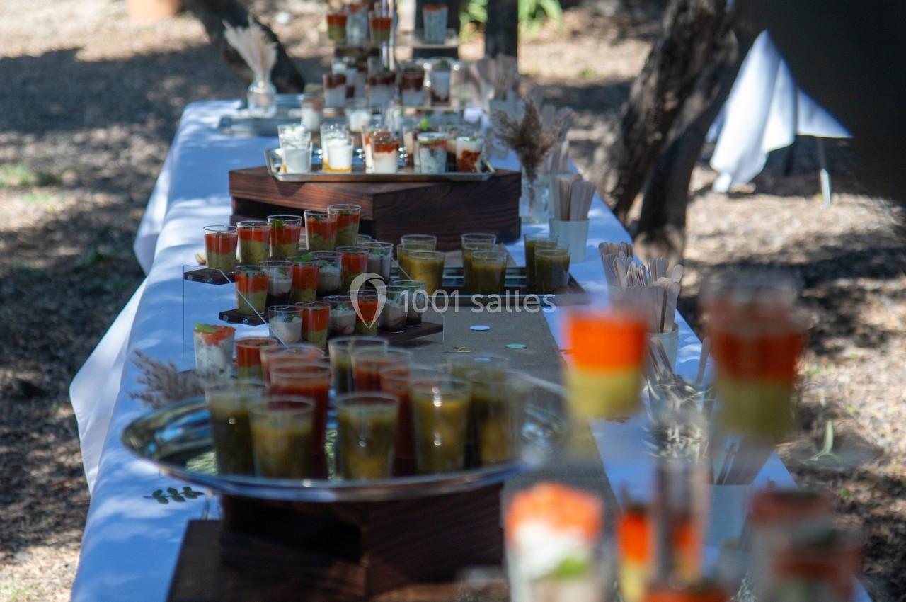 Buffet en plein air avec verrines colorées disposées sur une longue table blanche sous des arbres.