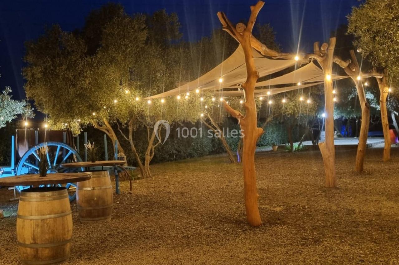 Espace extérieur éclairé par des guirlandes lumineuses, avec des tonneaux, un chariot en bois et des arbres.