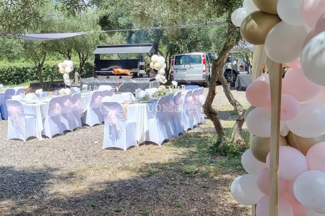 Tables décorées pour un événement en plein air, entourées d'arbres, avec un barbecue et des ballons en arrière-plan.