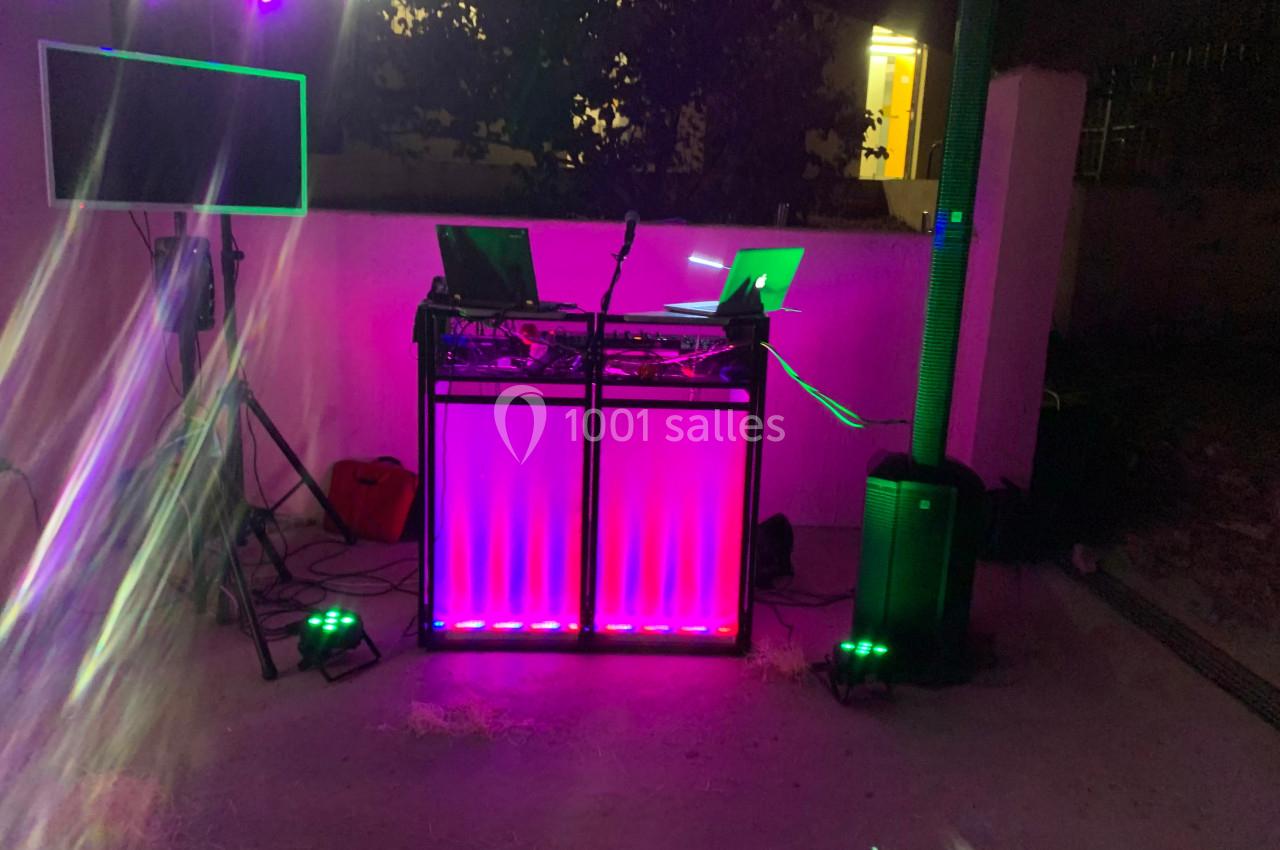 Poste de DJ éclairé par des lumières colorées, avec des équipements audio et un écran dans un espace extérieur sombre.