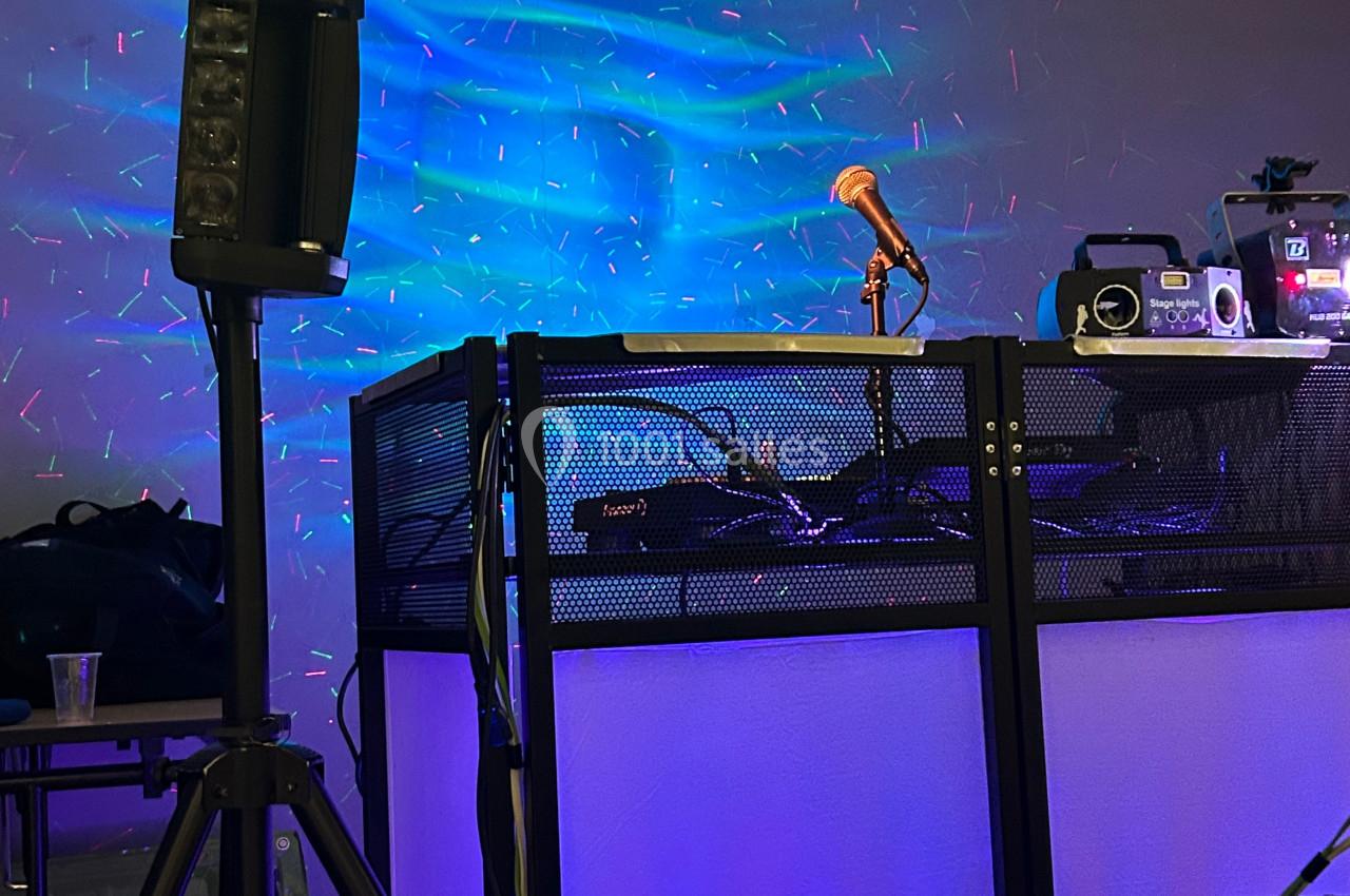 Un pupitre de DJ éclairé avec un micro orange, des équipements audio et des lumières colorées en arrière-plan.