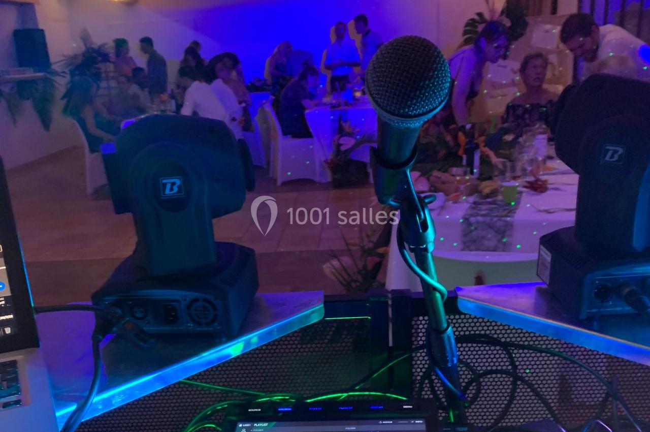 Vue depuis une table de DJ avec un microphone et des projecteurs, face à une salle éclairée en bleu avec des convives.